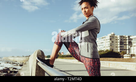 Active young woman stretching and warming up on the promenade along the ocean side. Female runner stretches along the seaside promenade. Stock Photo