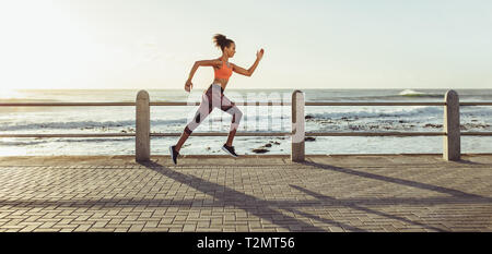 Athletic young woman running on seaside promenade. Side view of female runner sprinting outdoors. Stock Photo