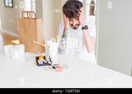 Young man eating asian sushi from home delivery suffering from headache desperate and stressed because pain and migraine. Hands on head. Stock Photo