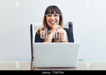 Close up of a smiling woman sitting at her desk in office with a laptop in front. Cheerful businesswoman in office working on laptop.