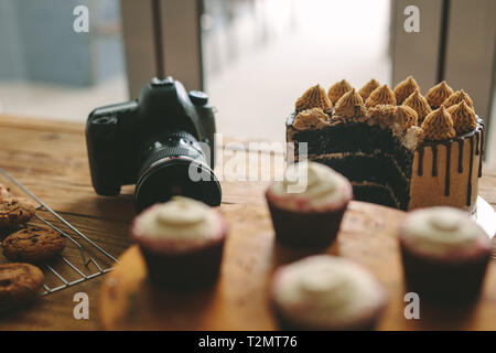 Digital camera on table with freshly made cakes, cookies and cupcakes on wooden table. Dslr camera with pastry items on a table. Stock Photo