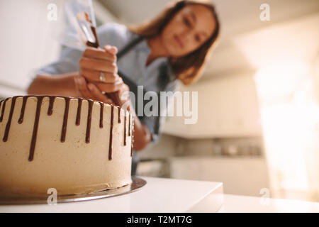 Cake on a wooden stand with chef decorating in kitchen. Baker decorating fresh delicious homemade cake with liquid chocolate on table. Stock Photo