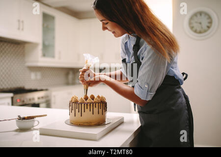 Female chef decorating cake with whipped cream using party bag. Woman in apron preparing a delicious cake at home. Stock Photo