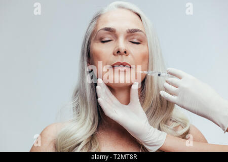 Senior woman having skin injections against grey background. Female getting anti aging shots on her face by beautician. Stock Photo