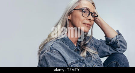 Senior caucasian woman sitting with hand on head and looking away thinking. Mature female with eyeglasses contemplating against grey background. Stock Photo