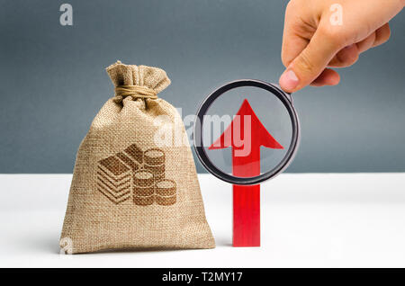 Male hand holds magnifying glass over money bag and up arrow. Concept analysis of profits and budget in the company. Successful business. The accumula Stock Photo