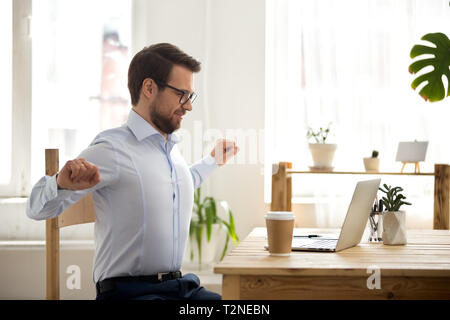 Happy satisfied businessman with eyeglasses sitting at the desk in office at workplace finish work, stretching out with raised hands. Millennial man r Stock Photo