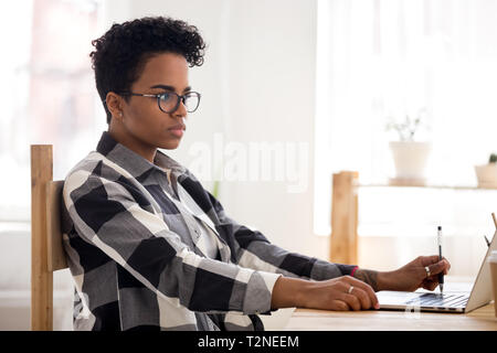 Concentrated african millennial woman with eyeglasses sitting at the desk in office room alone working using laptop. Young serious student or business Stock Photo