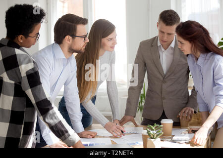 Five confident multiracial millennial diverse business people standing surrounding desk discussing in office boardroom. Young designers solving making Stock Photo