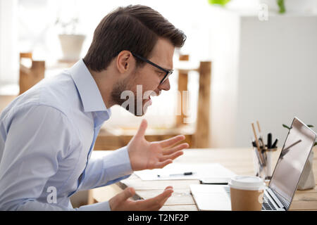 Irritated angry man sitting at the desk in office looking at laptop screen having problem, bad news. Side view of stressed, businessman noticed the er Stock Photo
