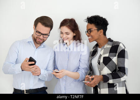 Group of people standing together opposite white wall looking on smartphone. Man showing to colleagues girls funny video or interesting news. Social n Stock Photo
