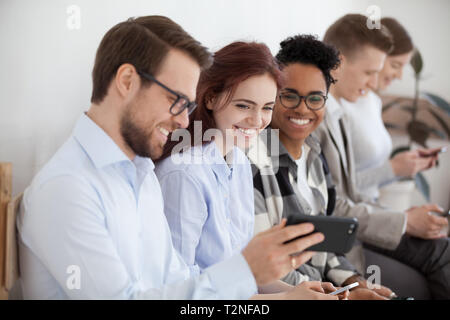 Diverse young businesspeople students friends sitting together in chairs in queue using mobile phones wait job interview or examination. Male showing Stock Photo