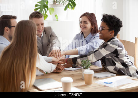 Excited happy group of young millennial businesspeople students stacking their hands palm, sitting in office at work. Gesture sign of support unity an Stock Photo