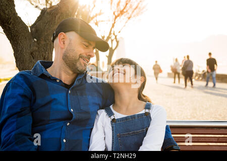 Father and daughter sitting on a bench on the waterfront looking at each other tenderly in a sunny day Stock Photo