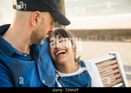 Father and daughter sitting on a bench on the waterfront having fun together in a sunny day Stock Photo