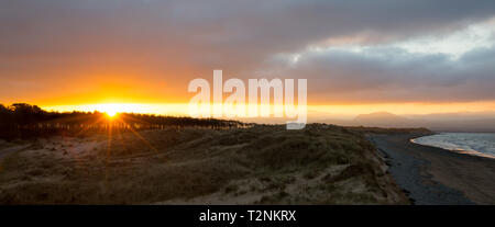 Panoramic view of sunrise at Newborough Warren, Anglesey. Sun rising over Snowdonia mountains in distance as dawn breaks; dark clouds, dark shoreline. Stock Photo