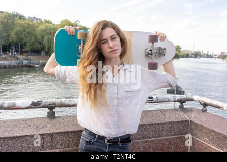 Young woman with skateboard on shoulder, river in background, Berlin, Germany Stock Photo