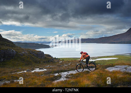 Male mountain biker biking down toward loch in mountain landscape,  Achnasheen, Scottish Highlands, Scotland
