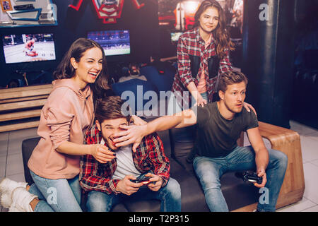 Two guys playing game on sofa in room. One of them hold hand in front of opponent's face. Young women laughing. They stand behind men Stock Photo