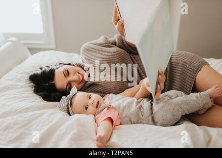 Mother reading with baby daughter on bed in bedroom Stock Photo