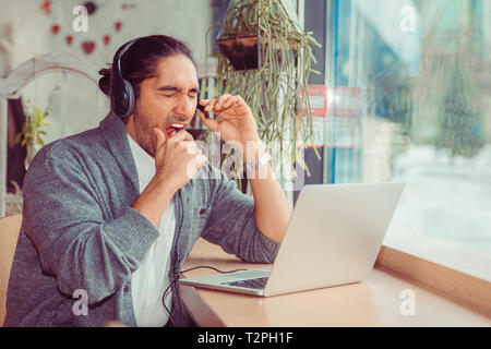 Handsome telemarketer man yawning. Closeup portrait of a handsome guy wearing casual clothing sitting near window at table in living room or coffee sh Stock Photo