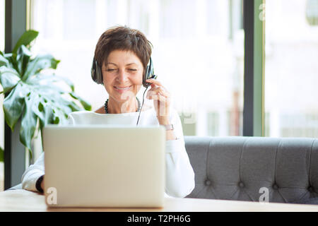Modern mature woman wearing, holding headset sitting at table in front of computer, laptop looking at it, smiling happily. Positive freelancer, custom Stock Photo