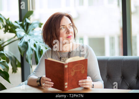Middle-aged mature elegant woman sitting at table in cafe with a book looking away thoughtful daydreaming. Stock Photo