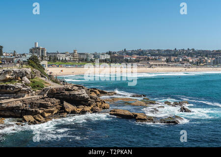 Sydney, Australia - February 11, 2019:  Sandy Bondi beach with gray rocky Mackenzies Point in front. Horizon is band of buildings of Bondi. Blue water Stock Photo