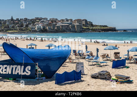 Sydney, Australia - February 11, 2019: Bondi Beach and North side cliffs with housing, under blue sky. Pale yellow sand with people, blue umbrellas an Stock Photo