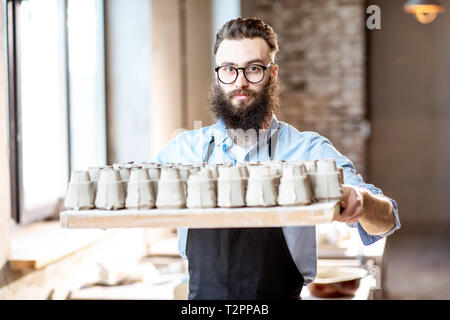 Portrait of a handsome bearded man standing with tray full of ceramic workpieces at the pottery shop Stock Photo