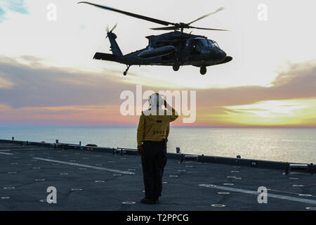 A U.S. Navy Sailor salutes a U.S. Army UH-60 Black Hawk helicopter and its crew members, assigned to the 1-108th Assault Helicopter Battalion, Kansas Army National Guard, as it takes off from a U.S. Navy ship in the Arabian Gulf, March 29, 2019.  The 1-108th AHB deployed to the Middle East to complete aviation missions in support of Operations Spartan Shield and Inherent Resolve.  (U.S. Army National Guard photo by Sgt. Emily Finn) Stock Photo