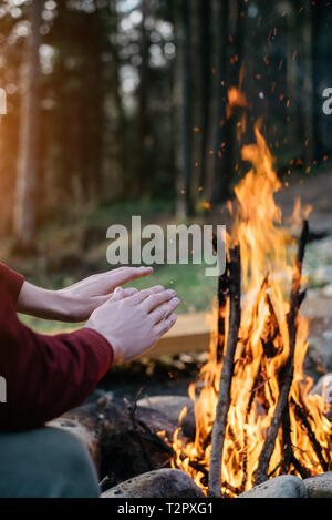 Outdoor vertical image of traveler warming his hands by near campfire an in forest. Rear view of traveler man in red jacket, sitting near to bonfire,  Stock Photo