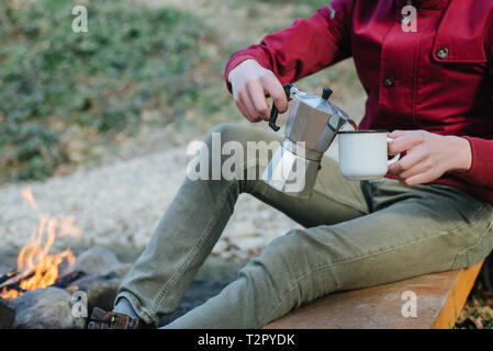 Young male pours itself hot beverage in mountains near to bonfire, relaxing after trekking. Traveler man in red jacket, sitting and holding a mug of c Stock Photo