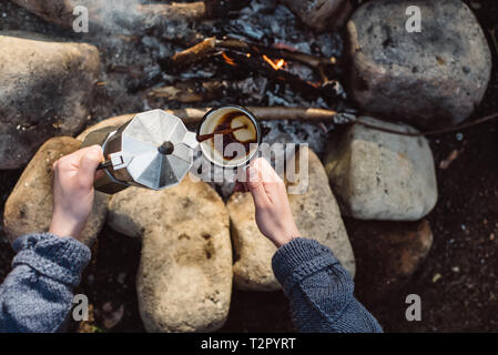View from above of traveler woman pours itself hot beverage in mountains near to bonfire. Female sitting and holding a mug of coffee after hiking. Tra Stock Photo