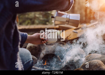 Traveler woman in blue sweater pours itself hot beverage in mountains near to bonfire, relaxing after trekking. Young female sitting and holding a mug Stock Photo