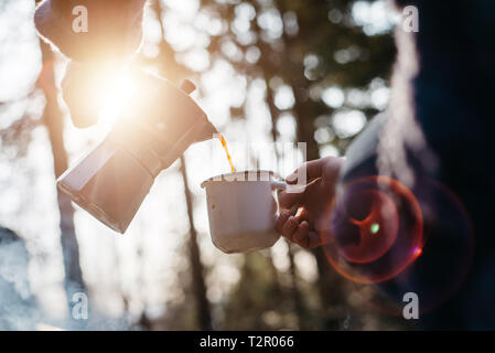 Outdoor shot of young woman pours itself hot beverage in mountains near to bonfire during the sunset. Traveler girl sitting and holding a mug of coffe Stock Photo