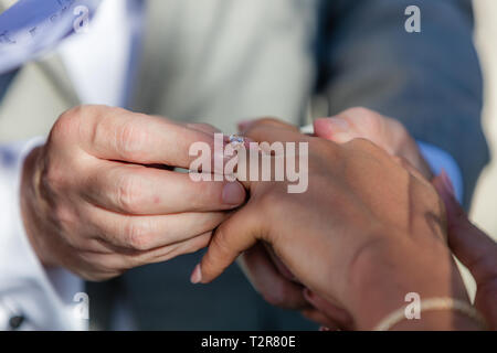 Groom putting a golden wedding ring on brides finger. Intermarriage - Caucasian man and Asian woman. Stock Photo