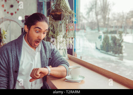 Man shocked looking to his wristwatch for time, her girlfriend being late to the date sitting in a coffee shop, or living room near window. Brunette e Stock Photo