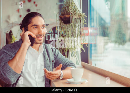 A man is talking on the phone and smiling. Closeup portrait of a handsome guy wearing formal white shirt and gray blouse sitting near window at a tabl Stock Photo