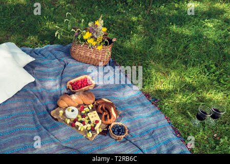 Basket with wine and flowers bread and cheese on a picnic blanket over green grass on the ground Selective focus Stock Photo