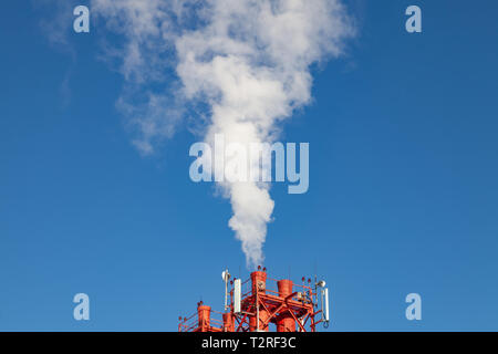 White harmful smoke coming out of the red-and-white pipes with mobile communication antennas at a factory in the city center against the background of Stock Photo