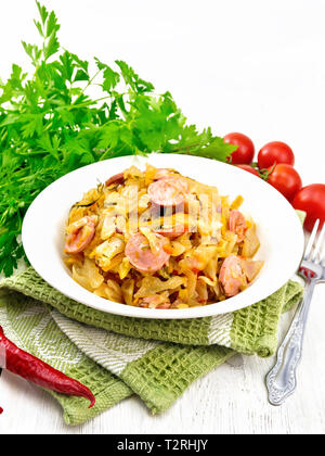 Cabbage stew with sausages in a white plate on a napkin, tomatoes, parsley and fork on light wooden board background Stock Photo