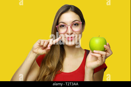 Portrait of a smiling young school nerd girl holding an apple gesturing like healthy good food isolated over yellow background Stock Photo