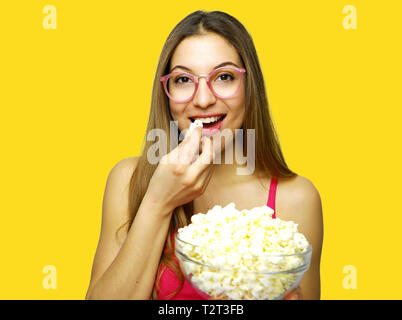 Happy cheerful woman eating pop corn on yellow background. Girl holding big glass bowl with cinema portion of pop corn. Stock Photo