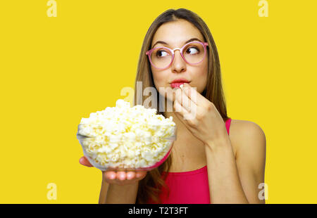 Girl eating pop corn and looking to the side on yellow background. Girl holding big glass bowl with cinema portion of pop corn. Stock Photo