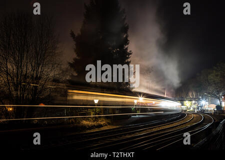 Atmospheric moving steam train & carriages travelling through vintage railway station at night in the dark. Fast movement motion blur. Concept speed. Stock Photo