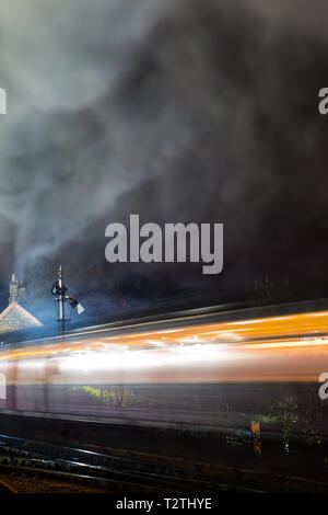 Atmospheric, moody night capture of moving UK steam train travelling through vintage railway station at night. Long exposure: movement, motion blur. Stock Photo