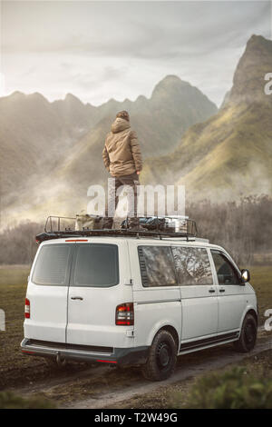 Man standing on the roof of a van surrounded by mountains and nature Stock Photo