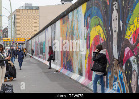 Tourists walking by, taking photos and posing in front of murals at the East Side Gallery, section of the Berlin Wall in Berlin, Germany. Stock Photo