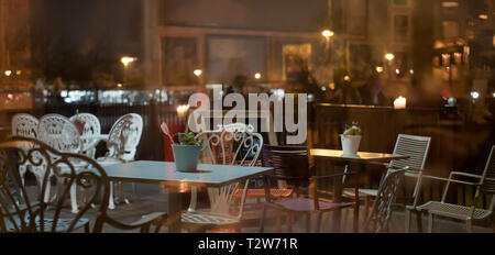 Interior, panoramic view of trendy bar/bistro/cafe taken from outside in the street in evening. Outside chairs & tables visible in window reflection. Stock Photo